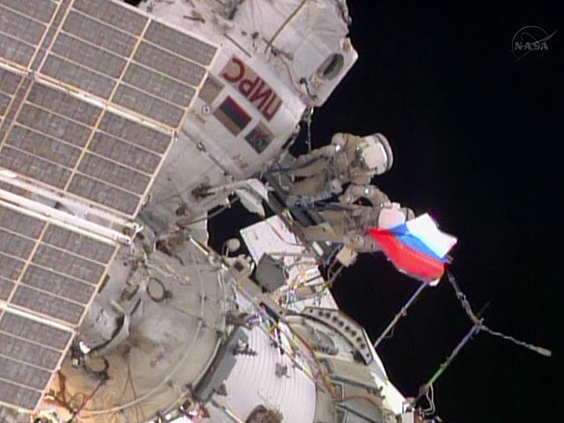 Engenheiros de voo Fyodor Yurchikhin e Alexander Misurkin seguram bandeira russa quase no fim da caminhada espacial  (Foto: Nasa TV/Reprodução)