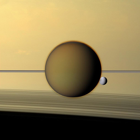A maior lua de Saturno, Titã, acompanhada de Dione, a terceira maior, com os famosos aneis do planeta ao fundo